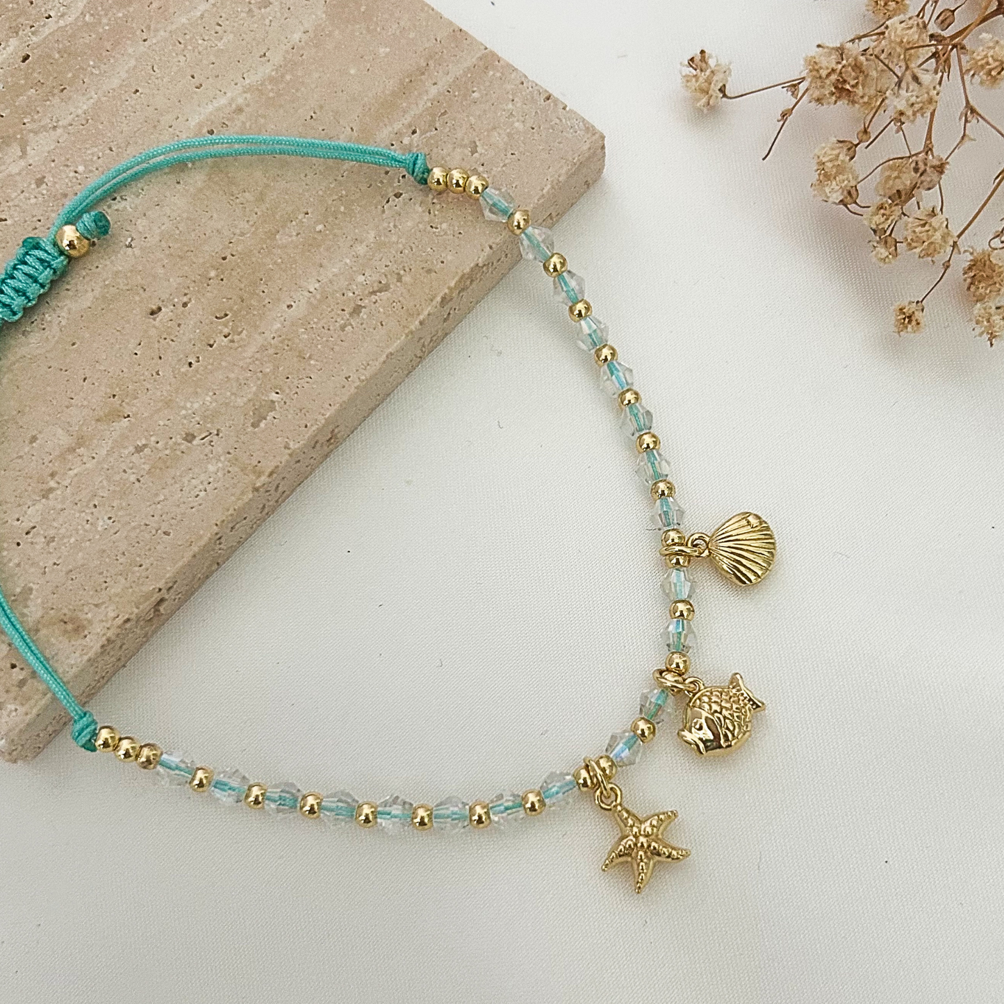 Delicate adjustable bracelet with shimmering crystal beads and gold sea-life charms, including a starfish, fish, and seashell, styled on a stone surface with dried flowers.
