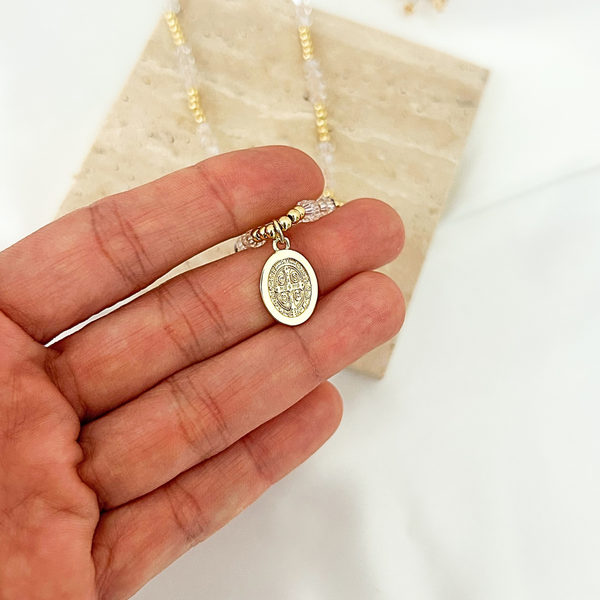 A gold Saint Benedict oval pendant necklace held in a person's hand, with a beaded chain and a cream stone display block in the background.