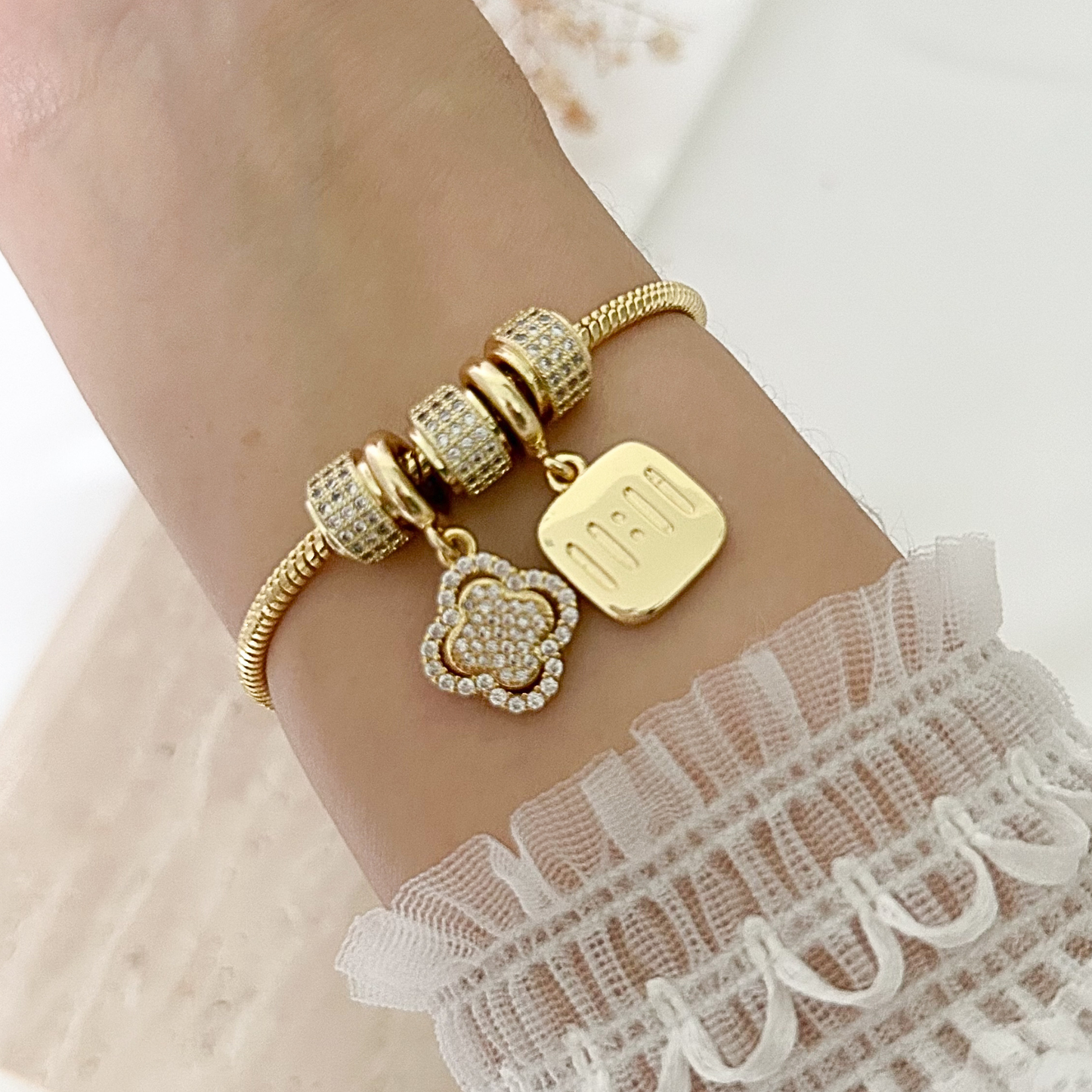 Gold-tone charm bracelet worn on the wrist, featuring sparkling pavé bead accents, a floral charm, and a square pendant engraved with “mom,” styled with a delicate white lace sleeve.