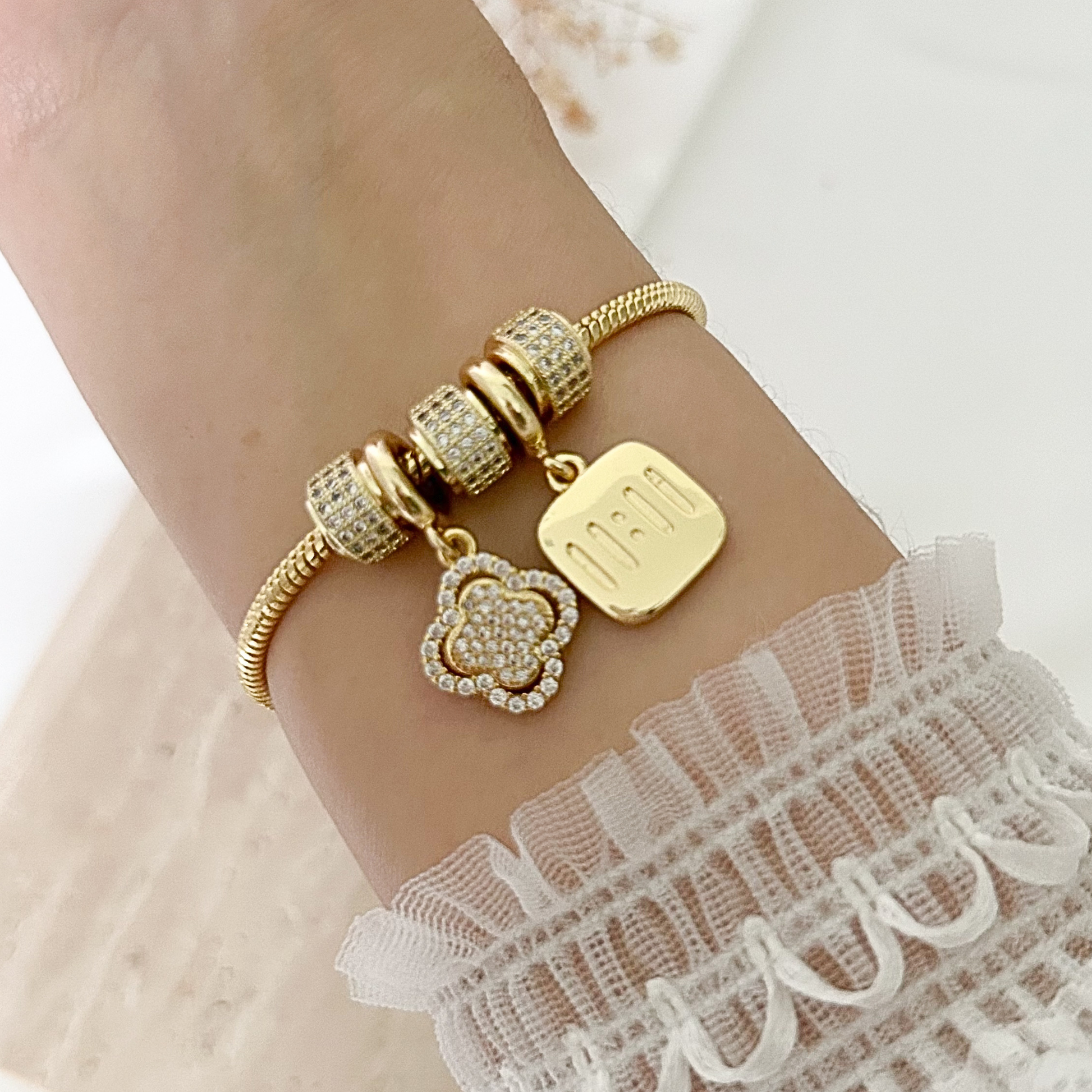 Gold-tone charm bracelet worn on the wrist, featuring sparkling pavé bead accents, a floral charm, and a square pendant engraved with “mom,” styled with a delicate white lace sleeve.