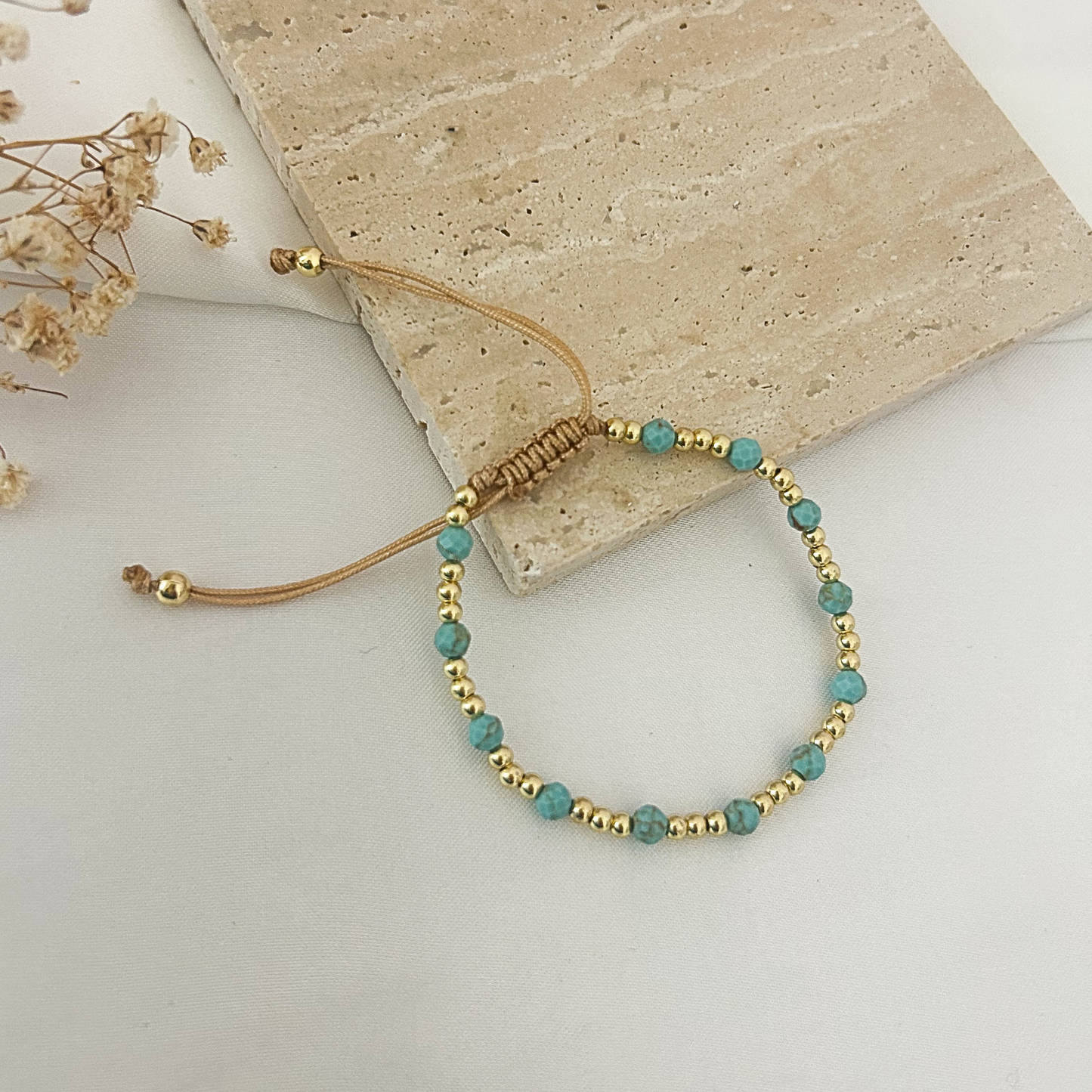 A beaded adjustable cord bracelet with turquoise-blue beads and small gold spacer beads, arranged on a light surface beside a beige stone tile and dried flowers.