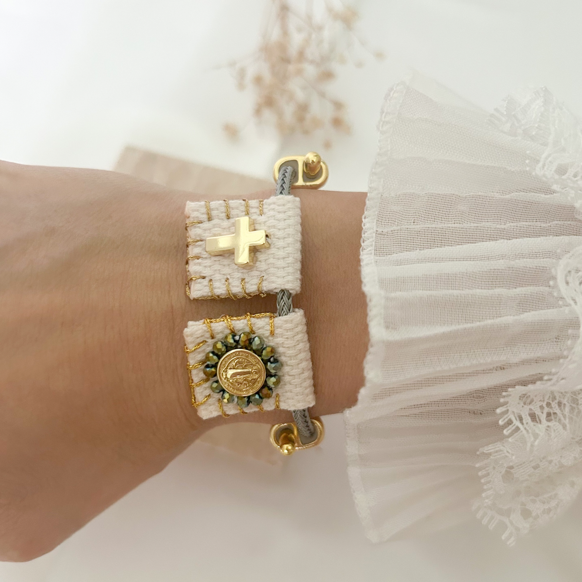 Handcrafted faith-inspired bracelet featuring a gold-tone cross and Saint Benedict medal on woven ivory fabric accents, displayed on a gray adjustable cord.