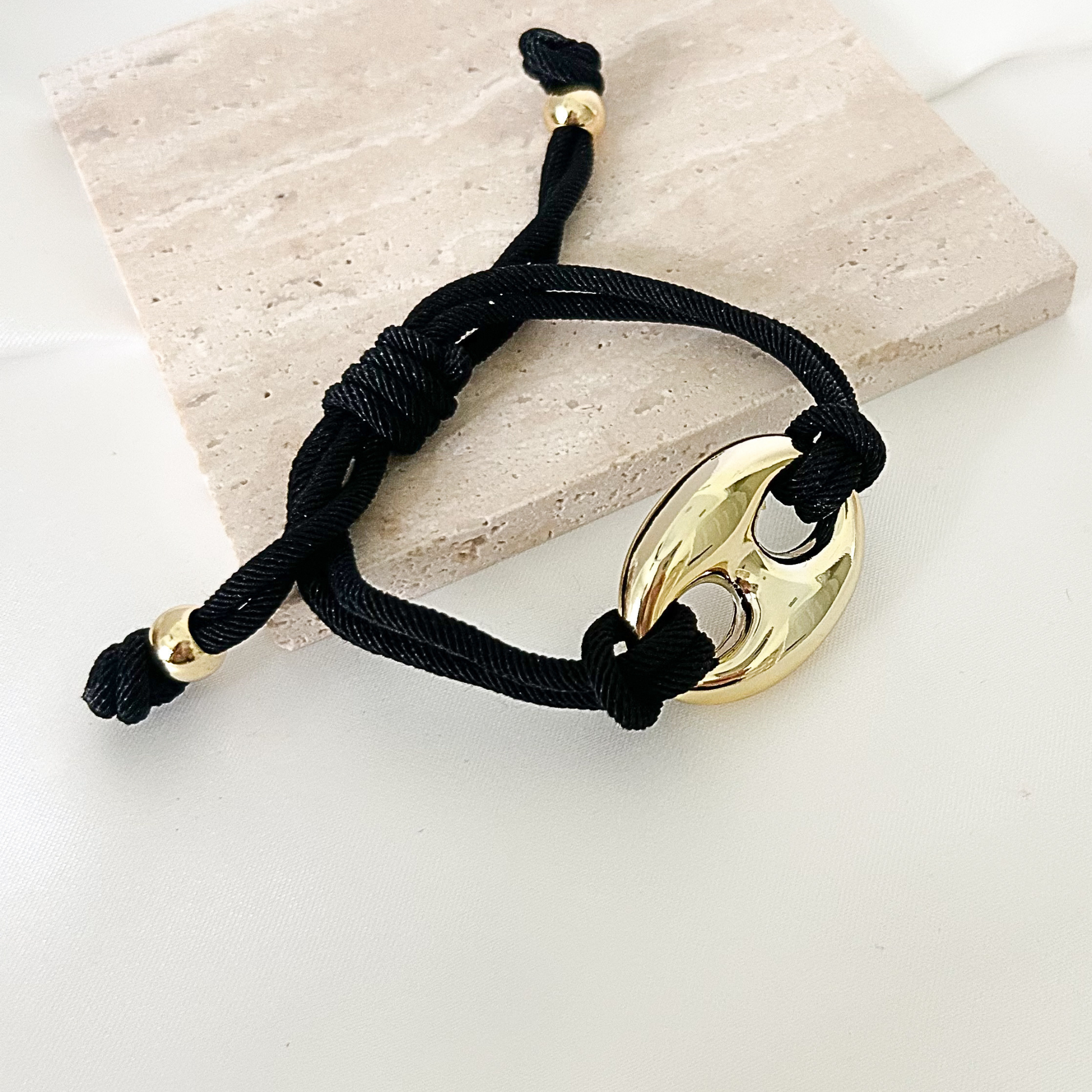 Minimalist black cord bracelet with a polished gold oval link centerpiece, adjustable sliding closure, and elegant modern design displayed on a beige stone surface.