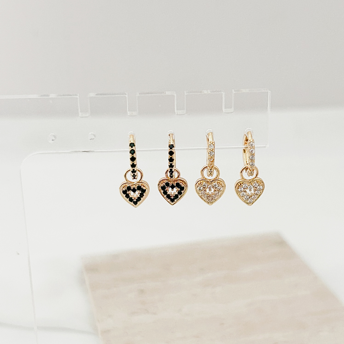 Four gold heart-shaped dangle earrings displayed on a clear acrylic stand against a white background. Two earrings feature black stones, and two feature clear pavé stones.