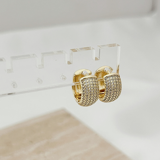 A pair of gold hoop earrings covered in small clear pavé stones, displayed on a clear acrylic stand against a soft white background.