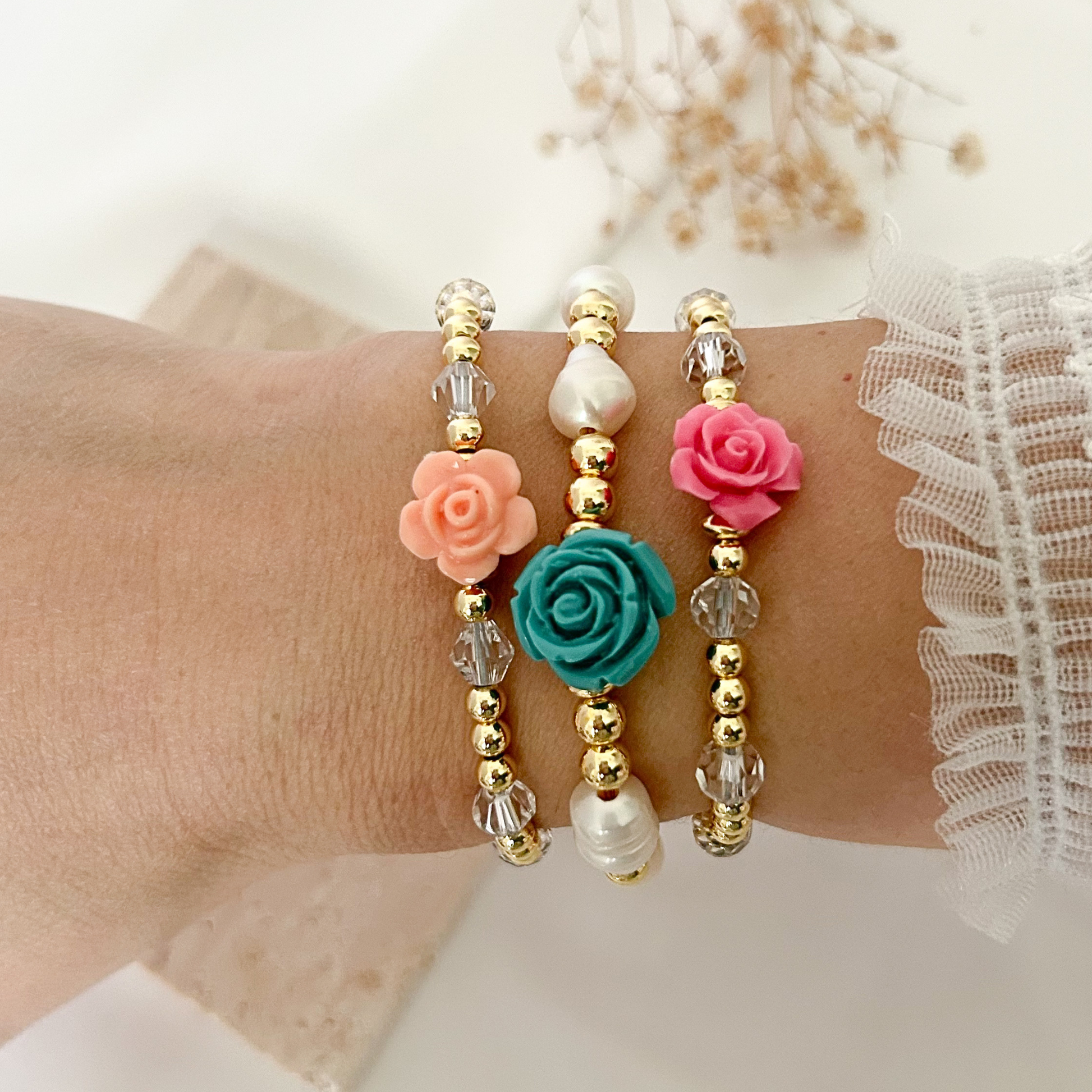 Set of three handmade floral bracelets featuring gold-tone beads, crystal accents, pearls, and resin roses in pink, peach, and turquoise, styled on a wrist against a soft neutral background.