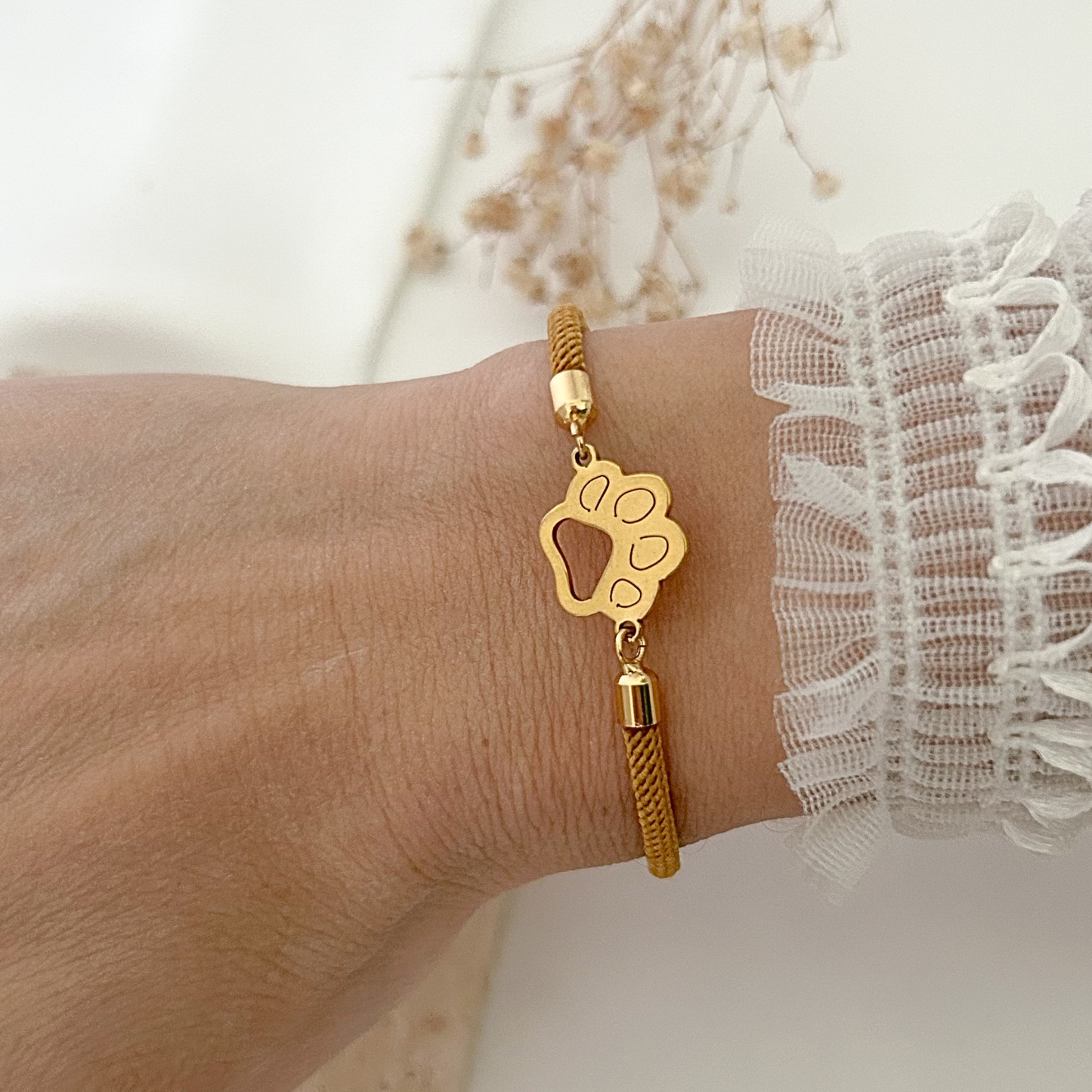 Close-up of an adjustable paw print bracelet worn on the wrist, combining a warm brown woven cord with polished gold-tone hardware for an elegant everyday look.