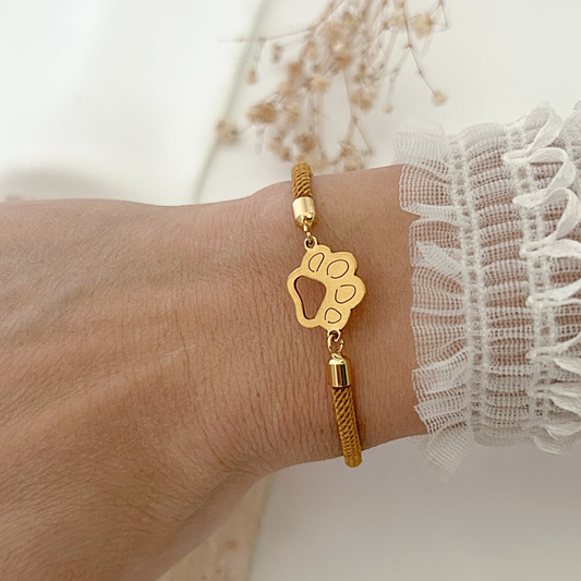 Close-up of an adjustable paw print bracelet worn on the wrist, combining a warm brown woven cord with polished gold-tone hardware for an elegant everyday look.