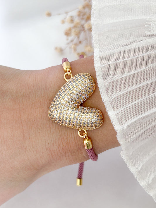 Gold heart-shaped bracelet with pink cord on a wrist against a white background
