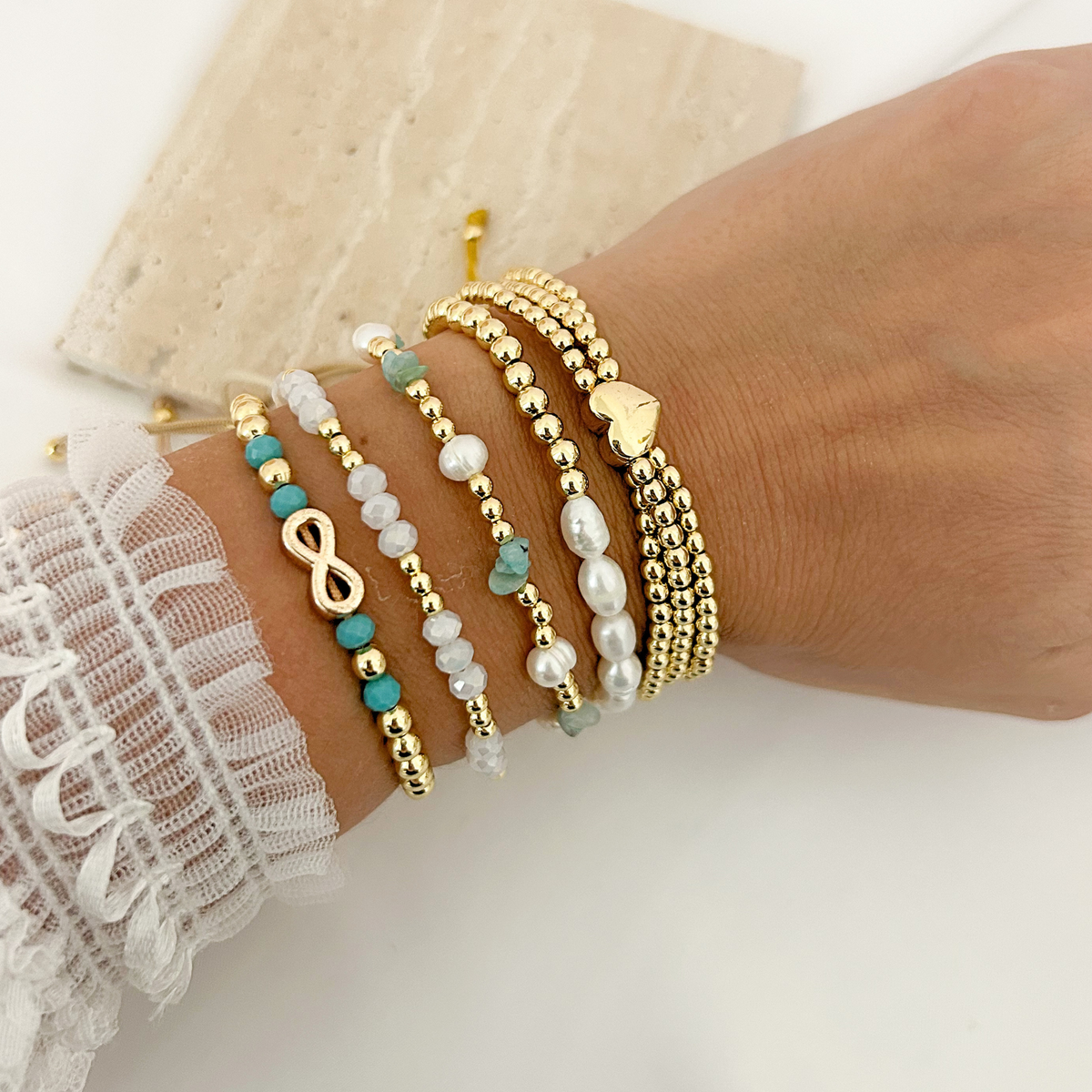 Gold beaded bracelet stack with turquoise accents, white crystals, freshwater pearls, an infinity symbol, and a heart charm.
