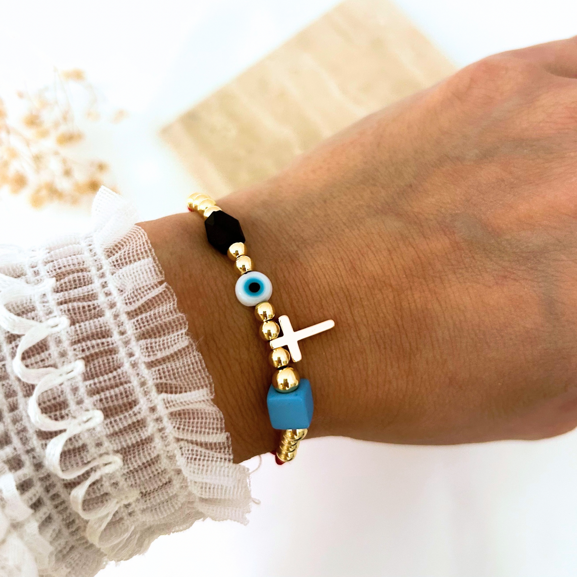 Adjustable red cord bracelet featuring gold-tone beads, a gold cross charm, a blue evil eye bead, and black and sky-blue accent beads. A meaningful everyday accessory that combines faith, protection, and modern style.