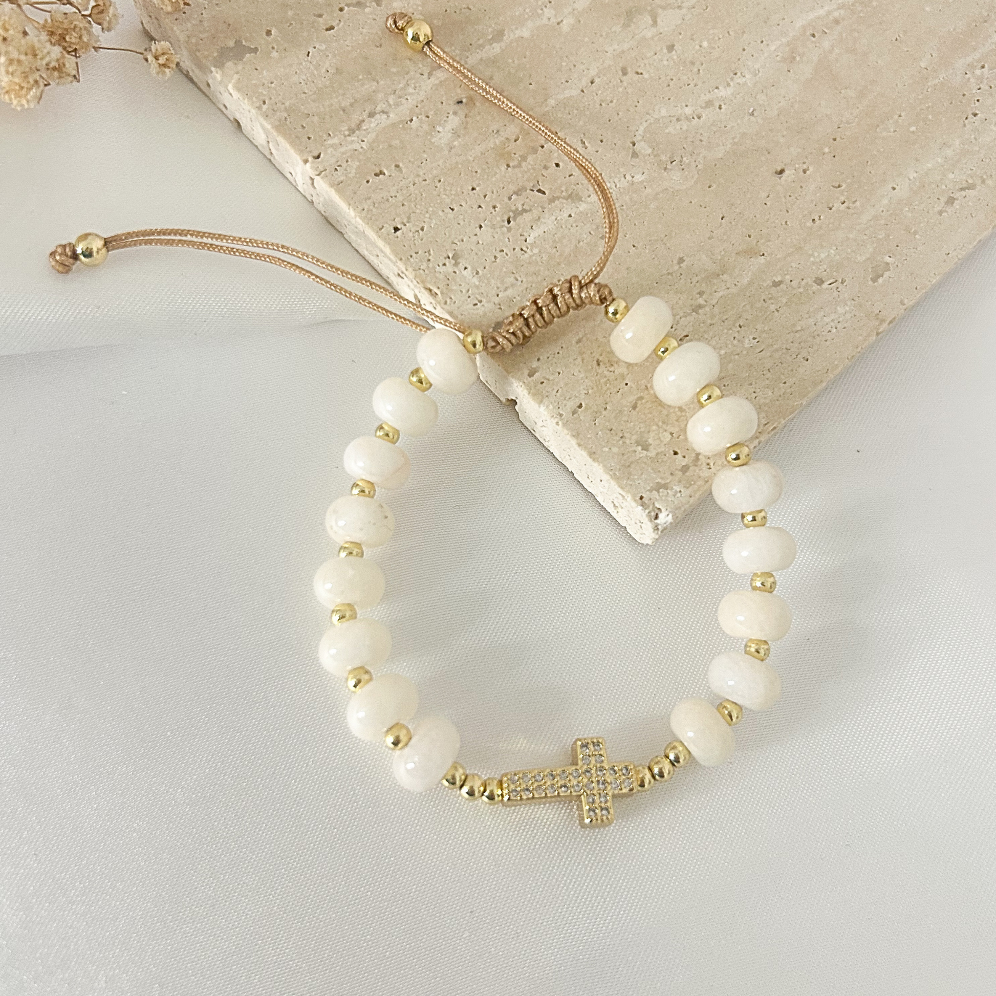 A beaded bracelet with white oval beads, small gold spacer beads, and a gold cross charm embellished with clear stones, displayed on a light surface next to a stone slab.