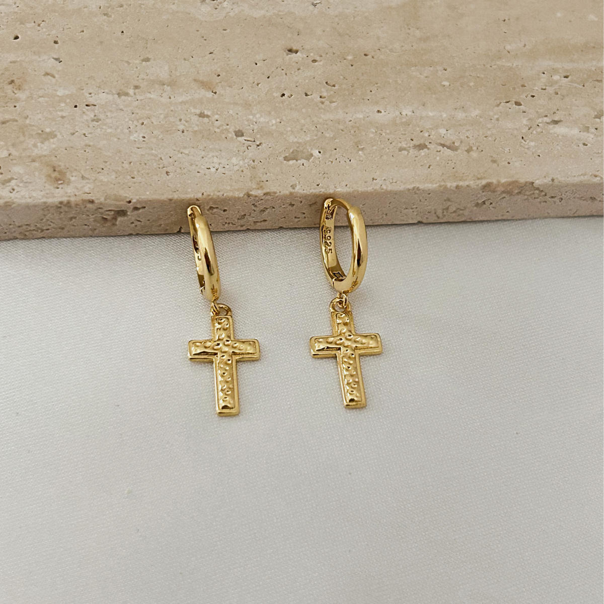 A pair of gold hoop earrings with small cross charms, displayed on a light gray surface with a beige stone slab in the background.