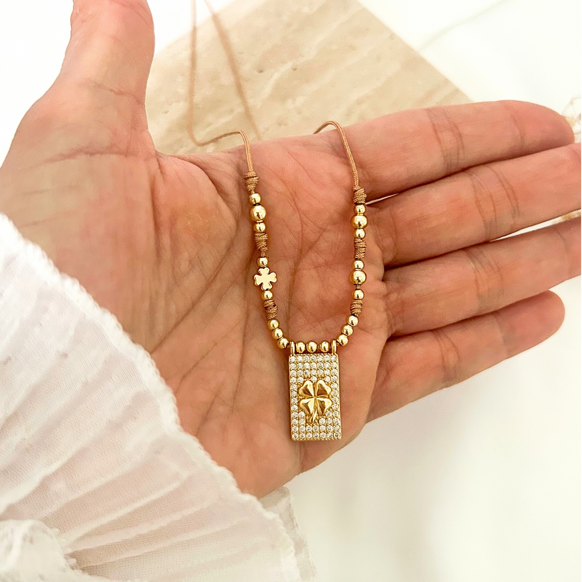 Gold-tone necklace with a crystal-encrusted rectangular pendant and raised four-leaf clover design, displayed on a neutral stone background with soft floral accents. A meaningful piece symbolizing luck and elegance.