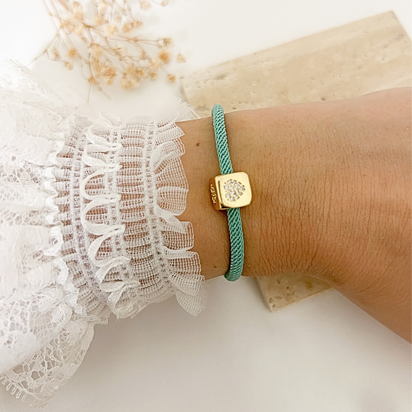 A close-up of a person's wrist wearing a mint green braided cord bracelet with a gold square charm featuring a clear crystal design. The person is wearing a white lace sleeve, and the background is a clean white surface with a decorative stone and dried flowers.