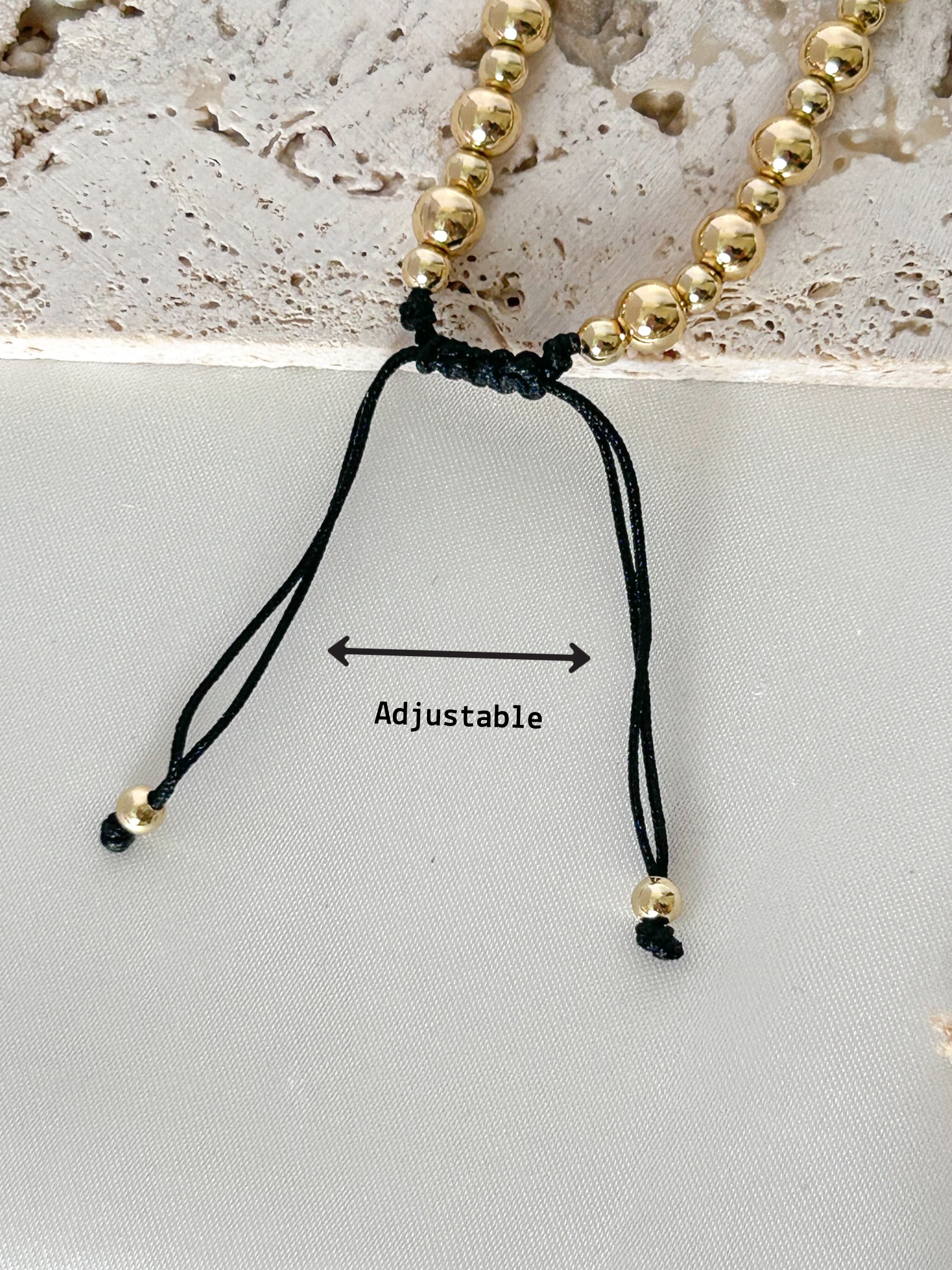 Gold beaded bracelet with adjustable black cord on a textured surface