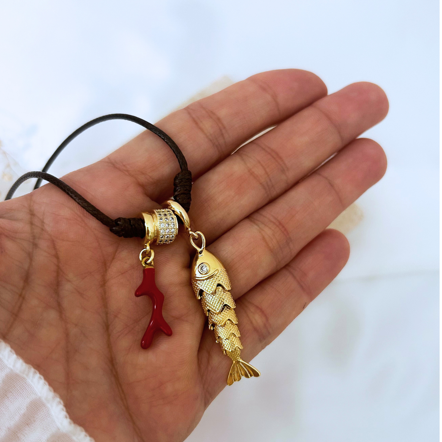 Close-up of a black adjustable cord necklace with a textured gold fish pendant, red coral charm, and crystal-accented gold bead. Lightweight, versatile, and perfect for everyday wear.