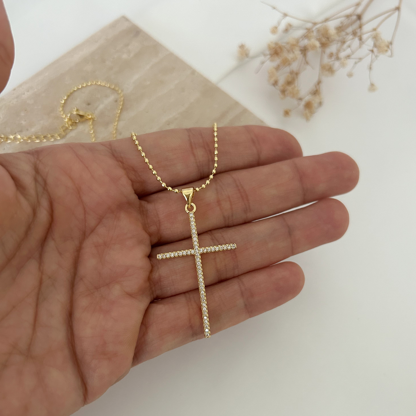 Mano sosteniendo un collar dorado con cruz de cristales y cadena tipo bolitas, resaltando los detalles brillantes de la pieza.