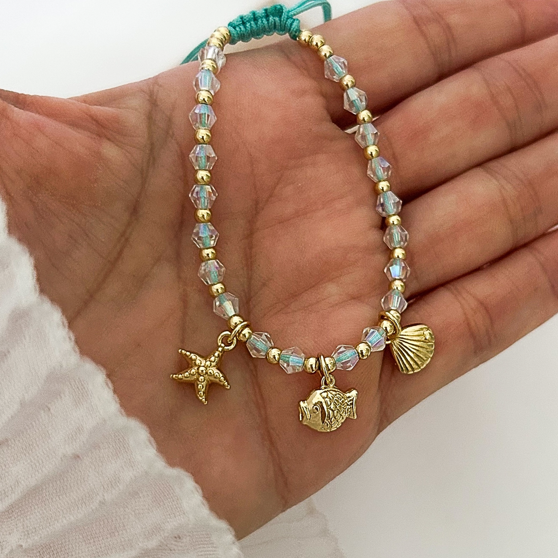 Close-up of a nautical-inspired bracelet showcasing iridescent crystal beads, gold-tone spacer beads, and dangling beach charms, photographed in hand against a soft neutral setting.