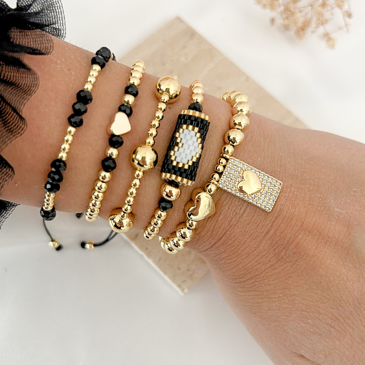 Black and gold bracelet stack featuring faceted beads, gold hearts, an evil eye design, and a sparkling rectangular charm.