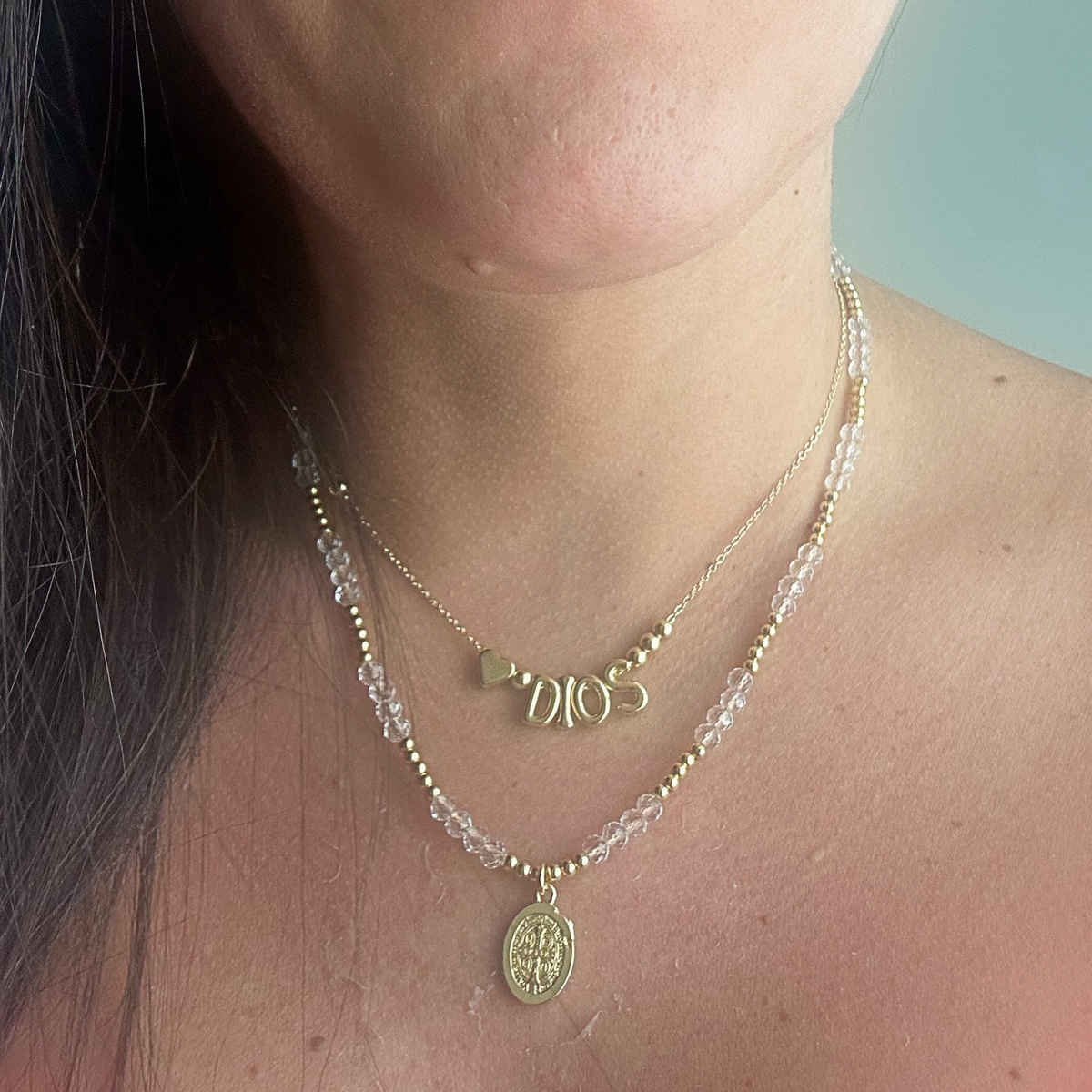 A close-up of a person's neck and chest wearing two layered gold necklaces. The shorter necklace features a 'DIOS' word charm, while the longer necklace has a clear faceted bead strand with a gold oval Saint Benedict medal pendant.