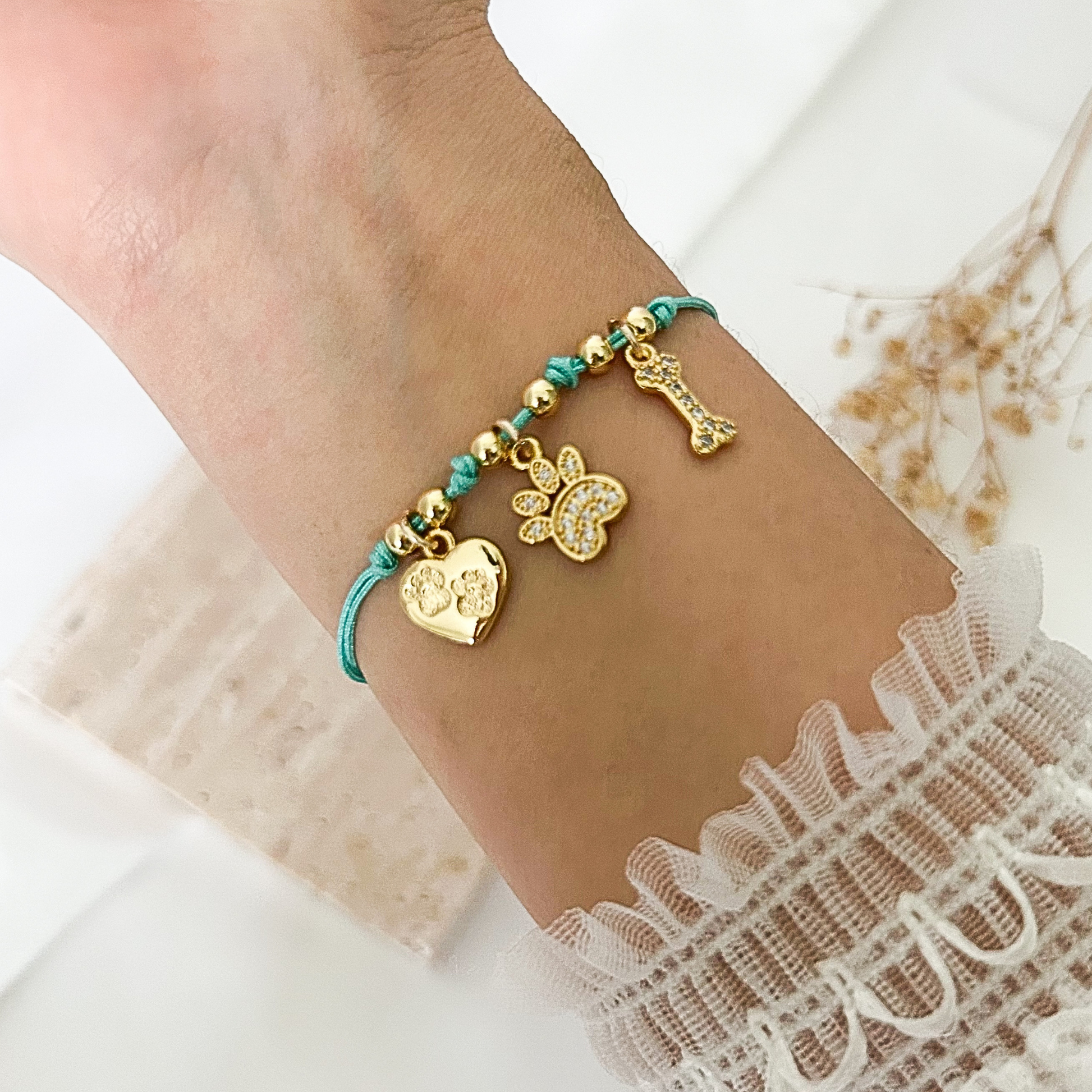 Adjustable pet lover bracelet with turquoise cord and gold-tone charms, featuring a heart with paw prints, a sparkling paw charm, and a crystal-studded bone charm, worn on the wrist with a delicate white lace sleeve.
