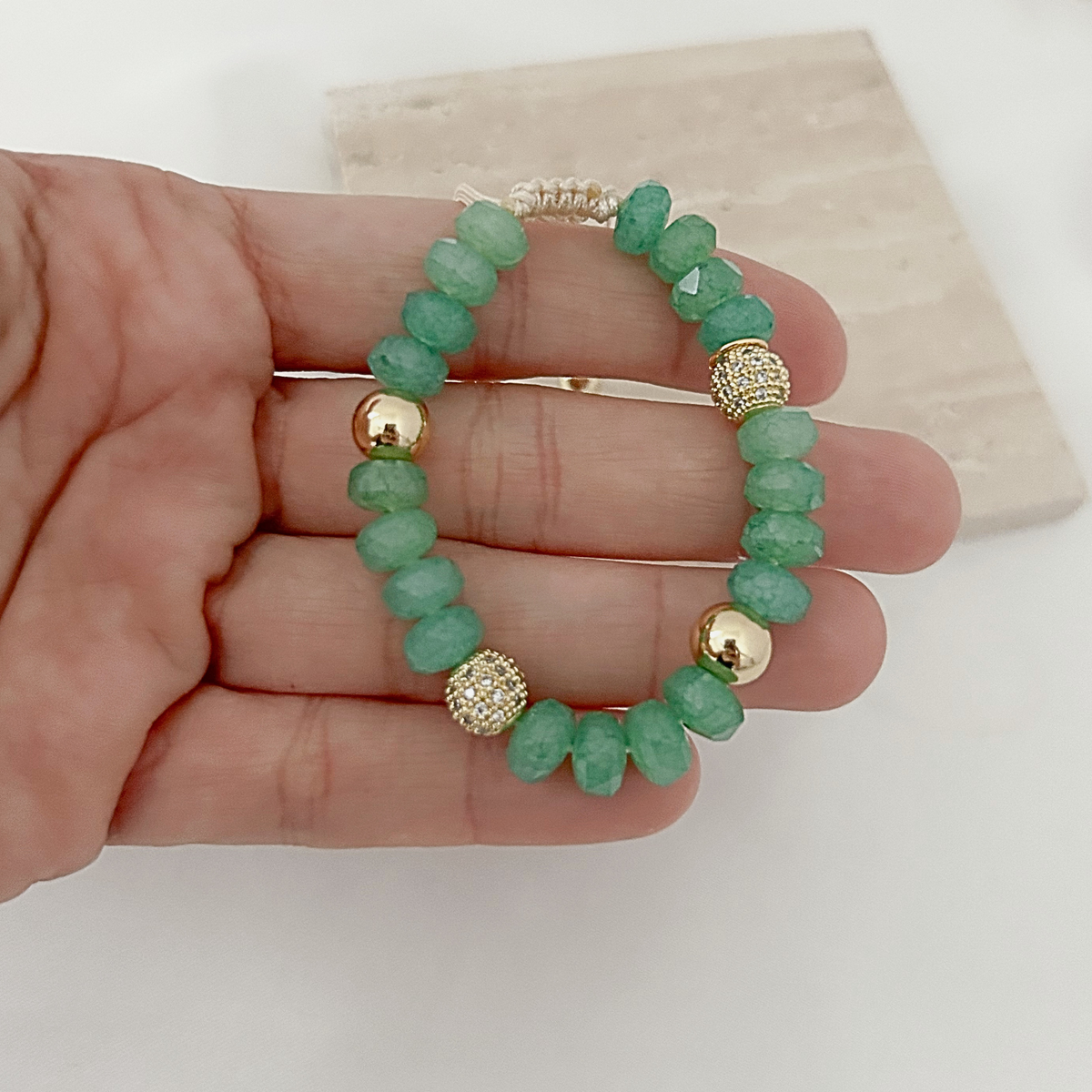 A hand holding a beaded stretch bracelet made of faceted green beads with gold-toned spacer accents, against a light neutral background.