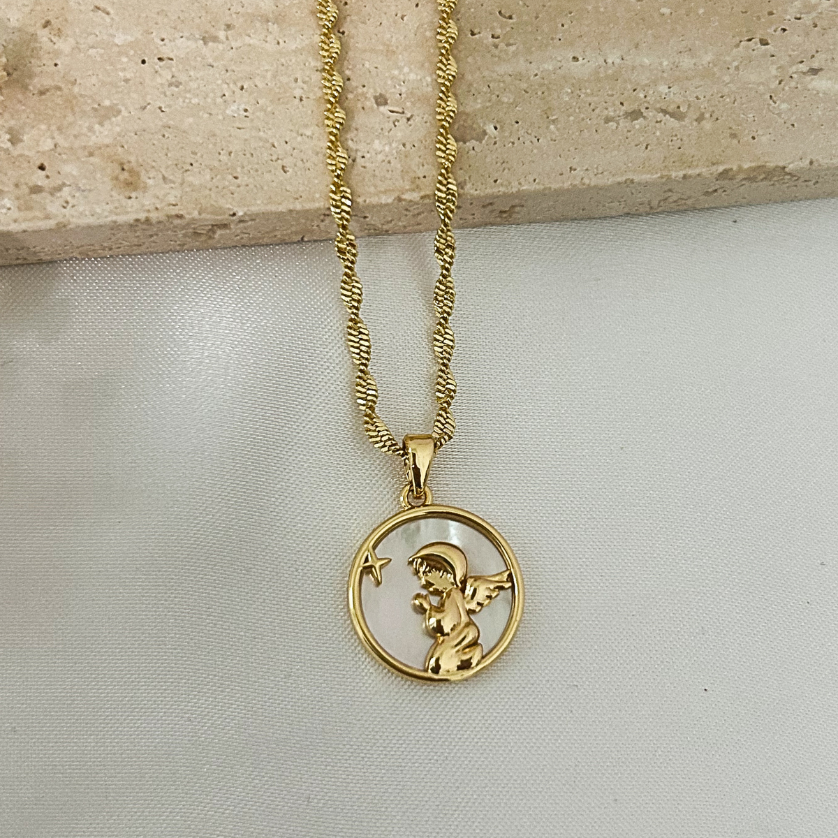 Close-up of a gold guardian angel pendant necklace featuring a mother-of-pearl background on a textured gold chain, photographed on a neutral minimalist background.