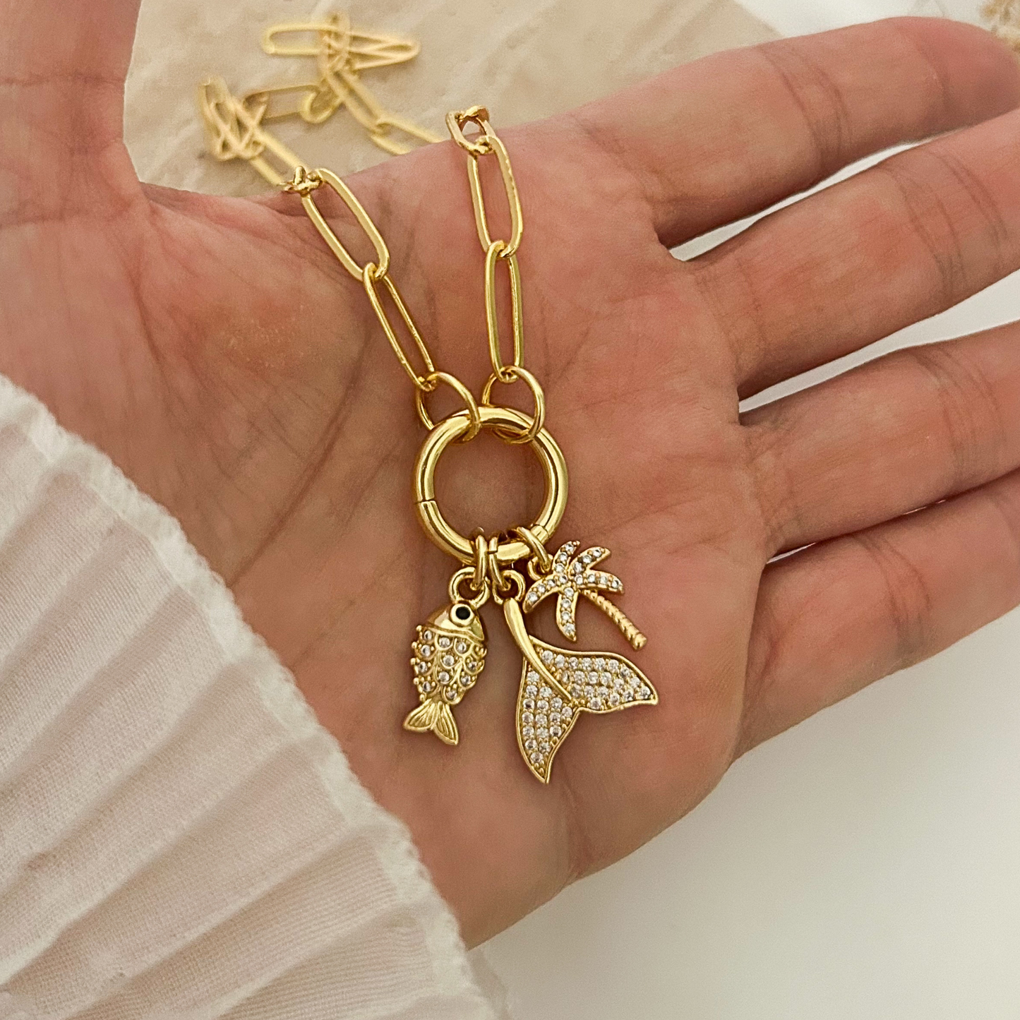 Gold paperclip chain necklace featuring three ocean-inspired charms: a sparkling fish, a crystal-studded mermaid tail, and a palm tree pendant, displayed in hand against a neutral background.