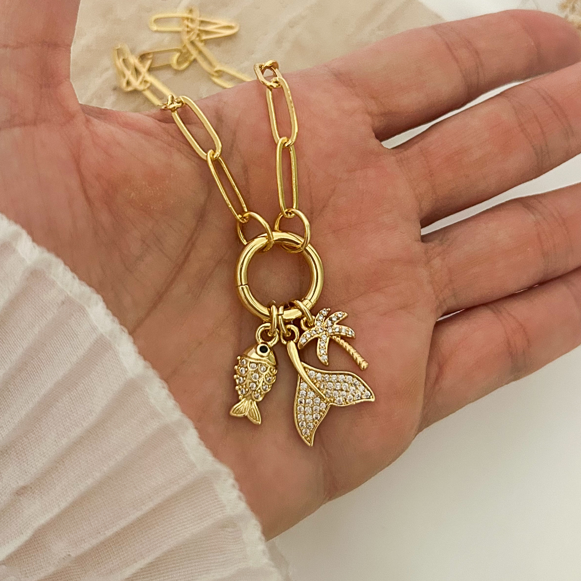 Gold paperclip chain necklace featuring three ocean-inspired charms: a sparkling fish, a crystal-studded mermaid tail, and a palm tree pendant, displayed in hand against a neutral background.