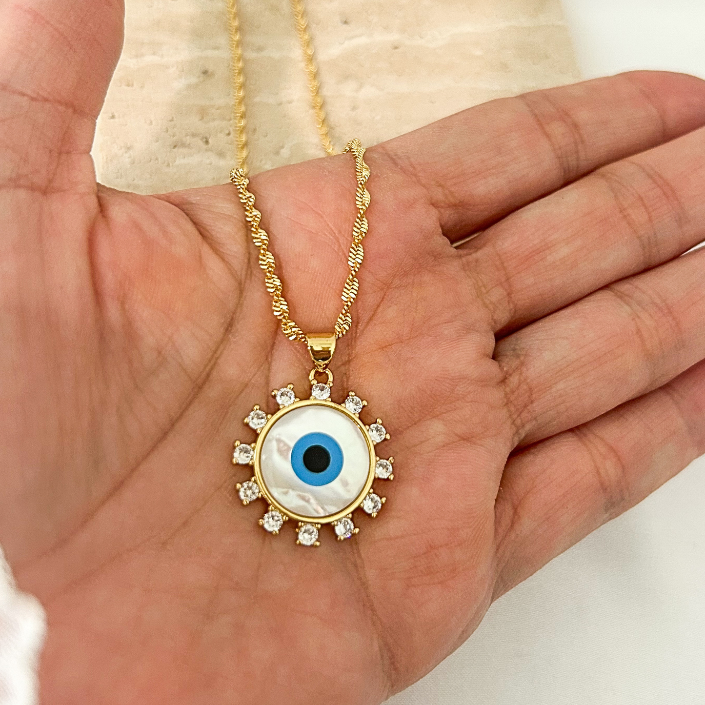 Gold evil eye pendant necklace with a mother-of-pearl center, blue enamel eye, and cubic zirconia halo, shown in hand to highlight its size, elegant details, and textured gold chai