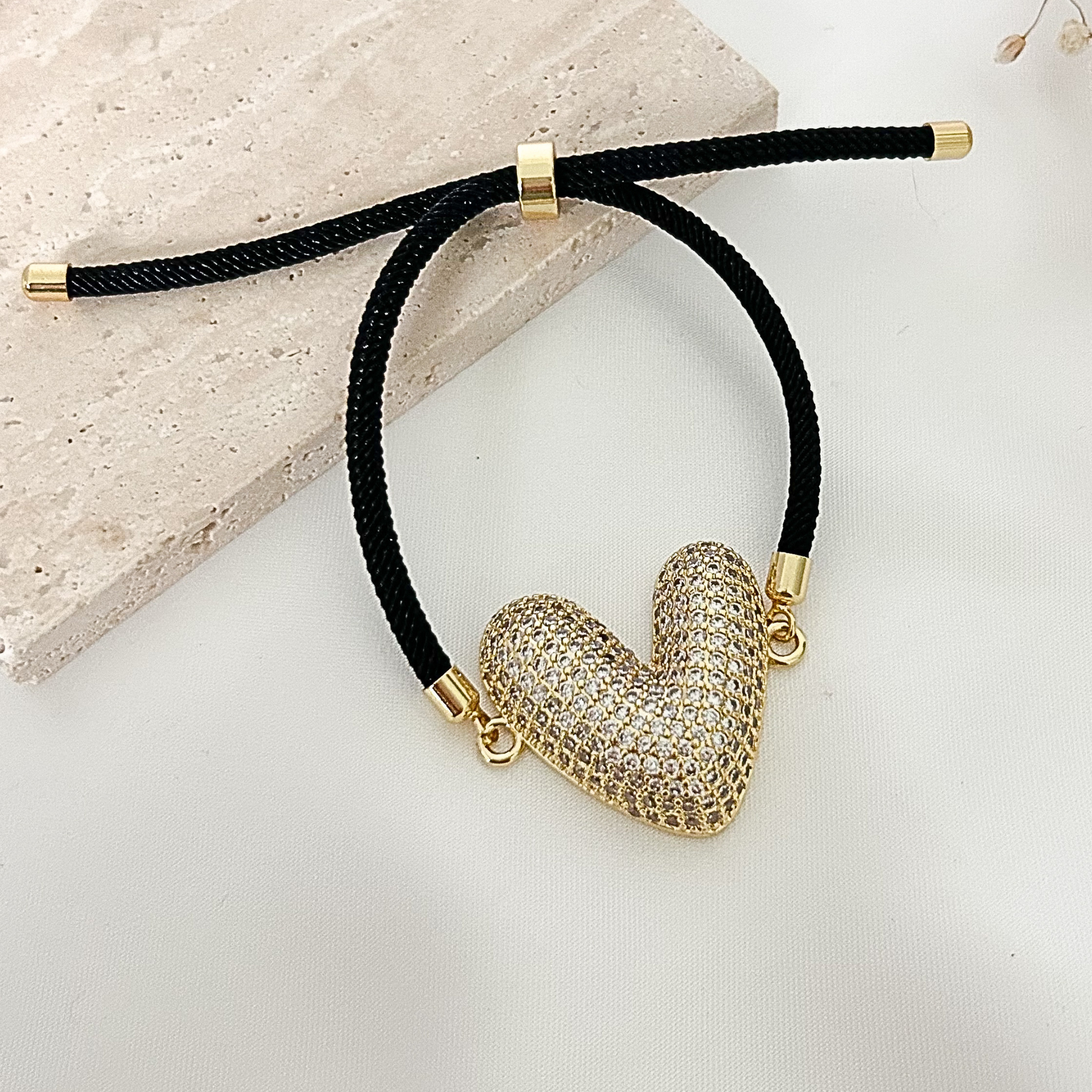 Adjustable black cord bracelet with a shimmering pavé heart charm, gold-tone accents, and a modern romantic design displayed on a neutral surface.