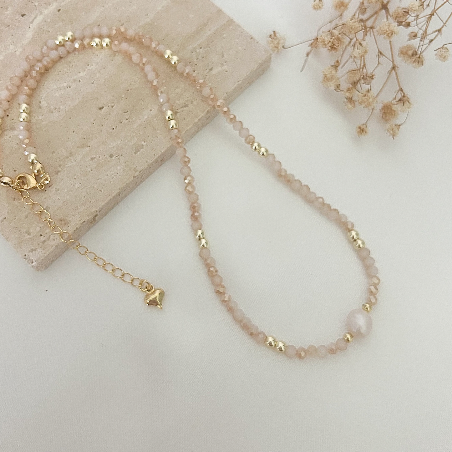 Handmade necklace with soft pink crystal beads, gold-tone spacers, and a natural freshwater pearl, displayed on a neutral stone background.