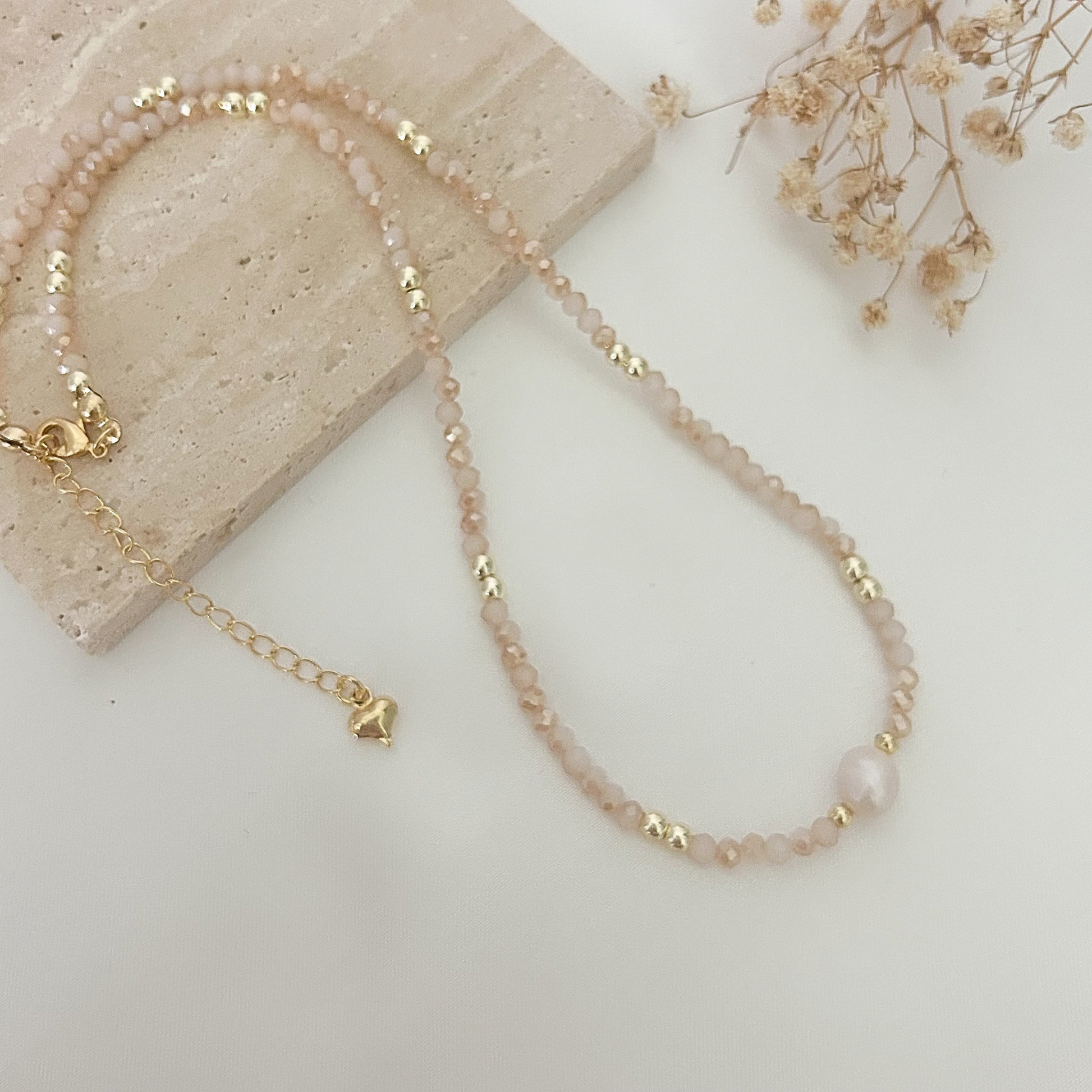 Handmade necklace with soft pink crystal beads, gold-tone spacers, and a natural freshwater pearl, displayed on a neutral stone background.