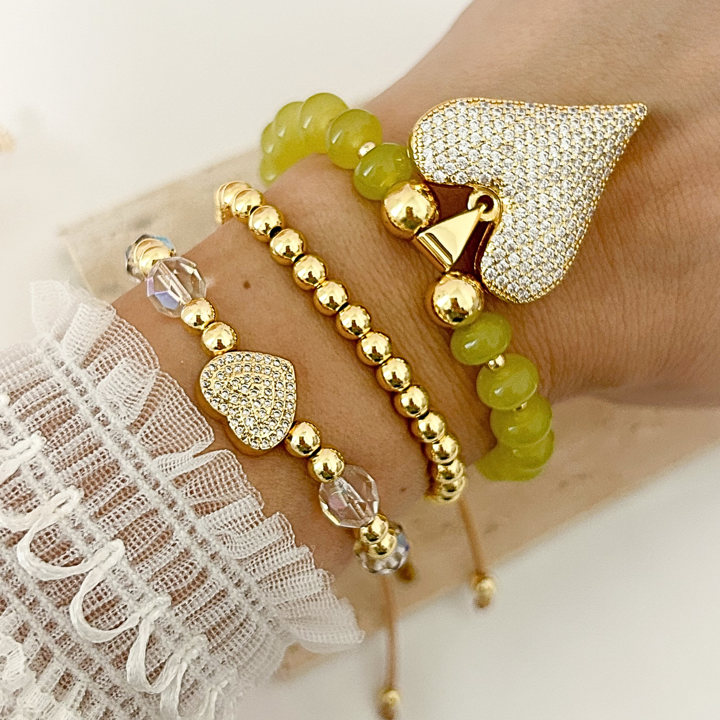 A close-up of a person's wrist wearing three beaded bracelets: one with translucent olive-green beads and a large pavé heart charm, one with gold-toned round beads, and one with gold-toned beads, clear crystal beads, and a pavé heart charm.