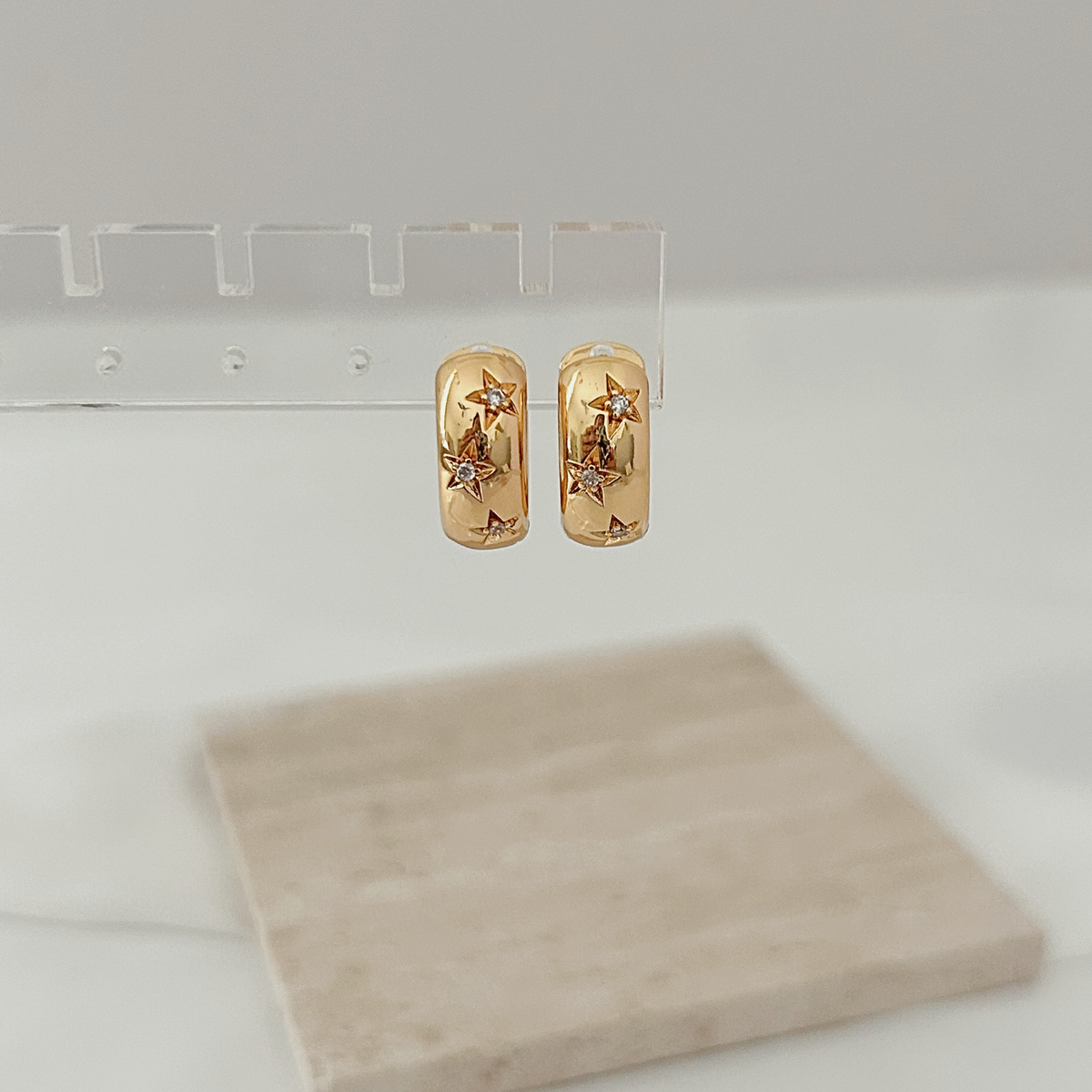 A pair of gold hoop earrings with star details, hanging from a clear acrylic display stand on a white surface with a blurred beige stone tile in the foreground.
