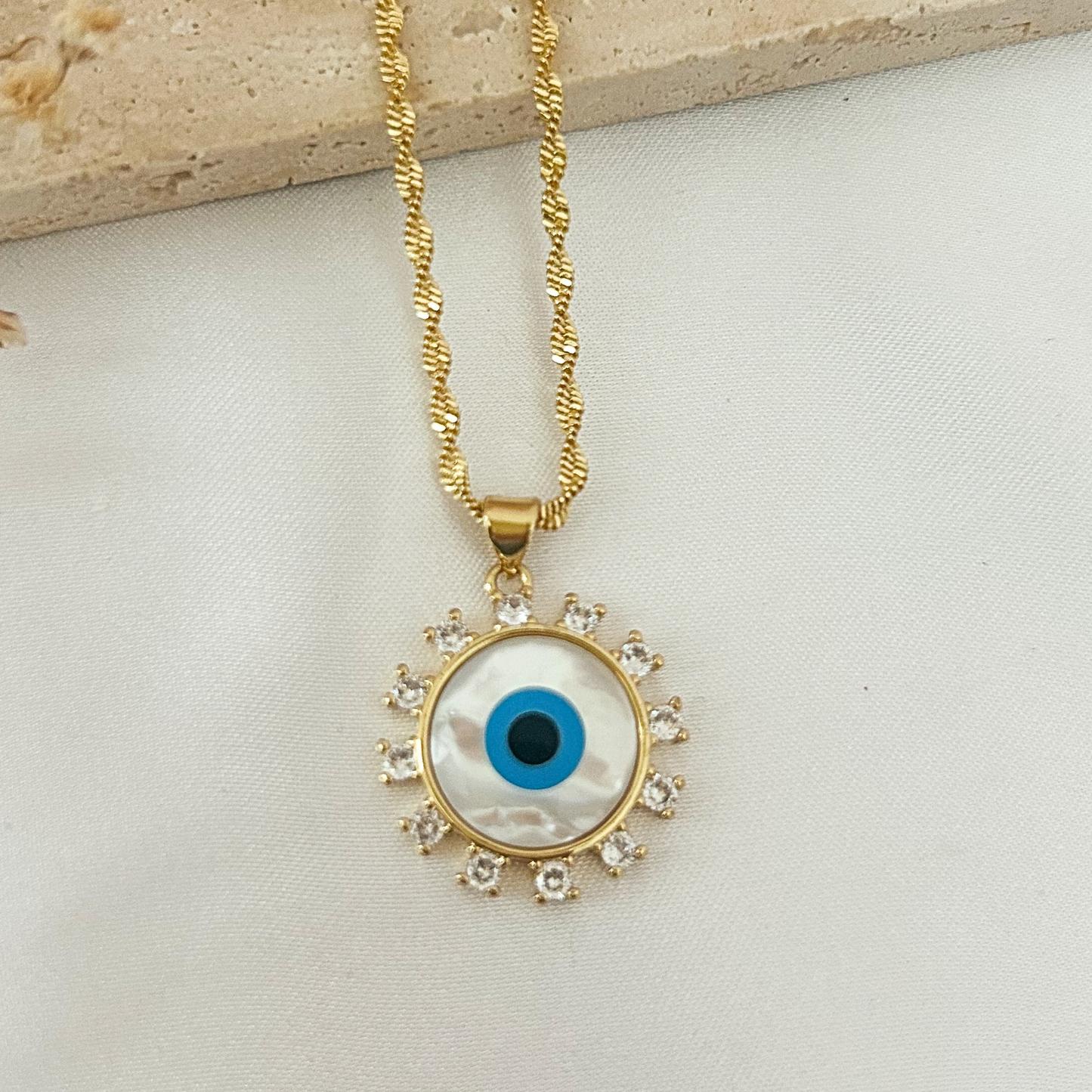 Close-up of a gold evil eye pendant necklace with a mother-of-pearl center, blue enamel eye, and sparkling cubic zirconia halo on a textured gold chain.