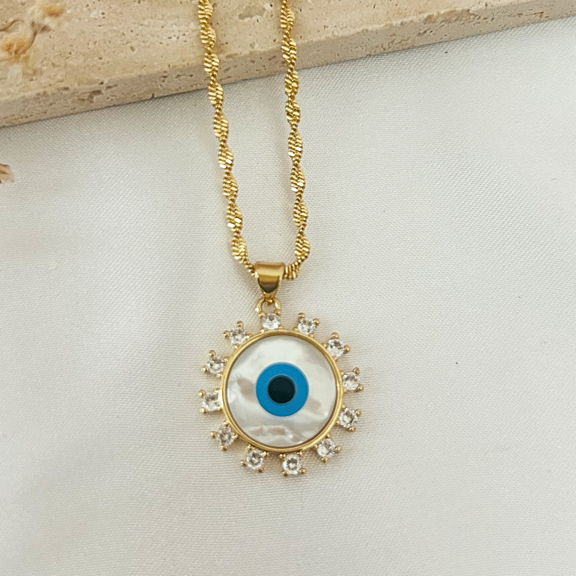 Close-up of a gold evil eye pendant necklace with a mother-of-pearl center, blue enamel eye, and sparkling cubic zirconia halo on a textured gold chain.