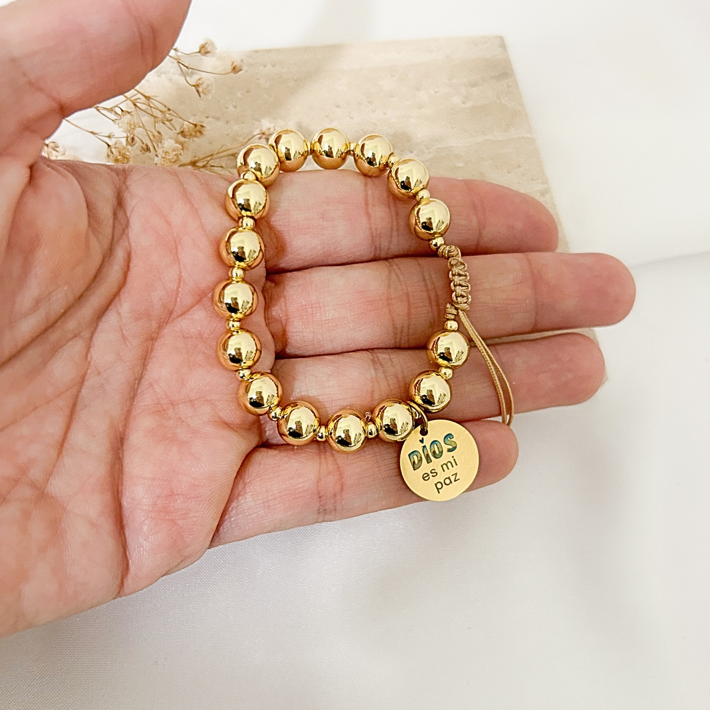 A gold beaded bracelet with a round charm engraved 'DIOS ESTÁ MÍ' is displayed on a person's hand against a light background.