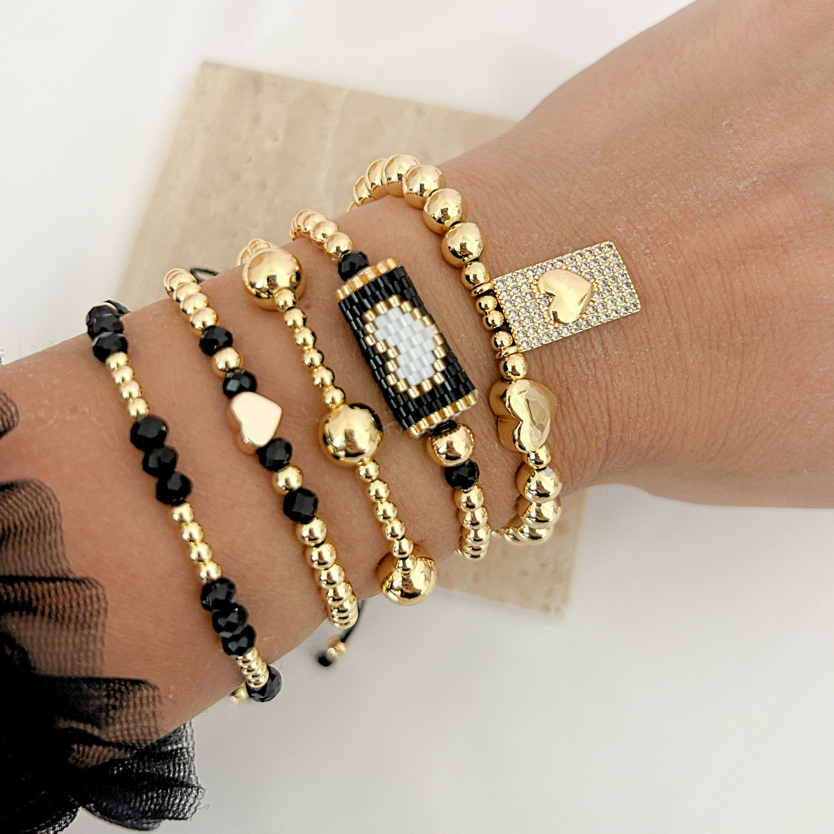 Close-up of layered black and gold beaded bracelets with heart charms, an evil eye centerpiece, and crystal details.