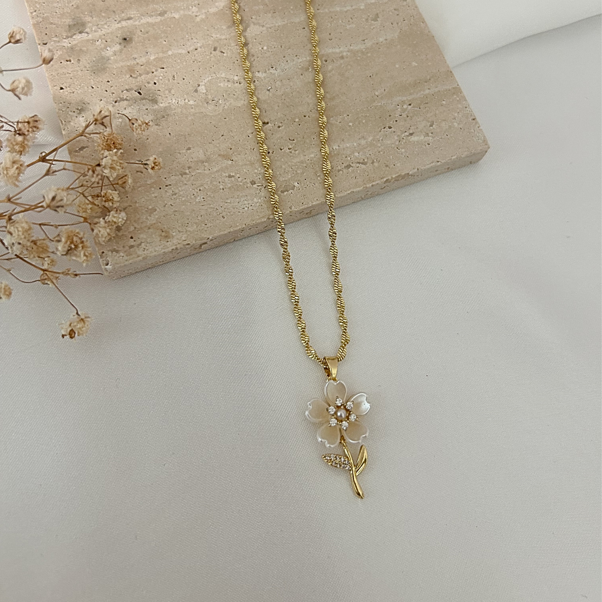 Gold flower pendant necklace featuring mother-of-pearl petals, pearl accents, and sparkling cubic zirconia details on a textured gold chain, displayed on a neutral minimalist background.