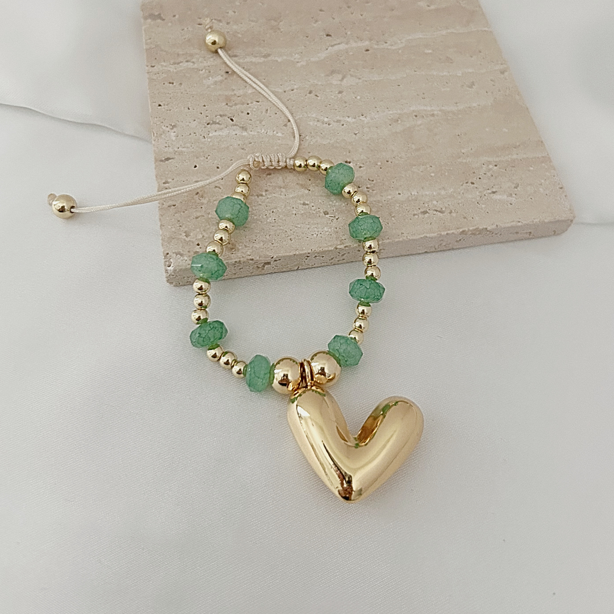 A gold-plated adjustable cord bracelet with green faceted beads and a large polished gold heart pendant, displayed on a light beige stone tile over a white surface.