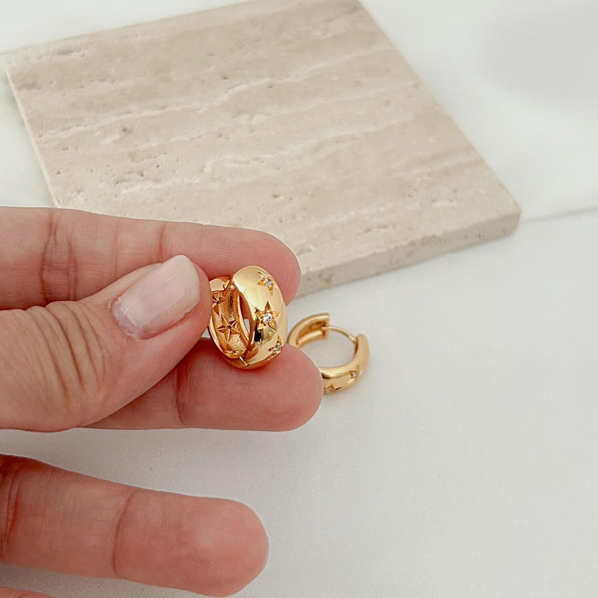 A close-up of a hand holding a small gold huggie hoop earring with star details, with another matching earring resting nearby on a white surface and a beige stone tile in the background.