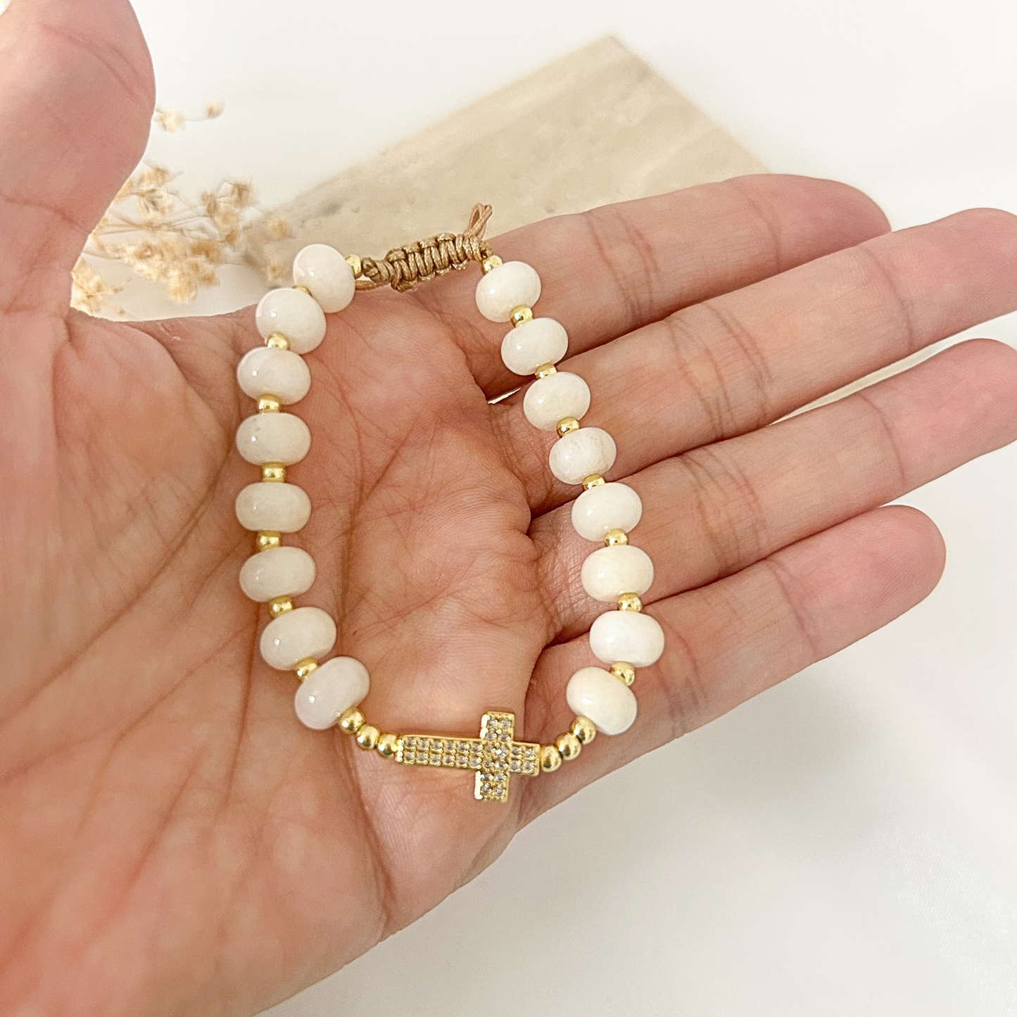 A beaded bracelet with white oval beads, gold spacer beads, and a gold cross charm embellished with small clear stones, shown resting on an open hand.