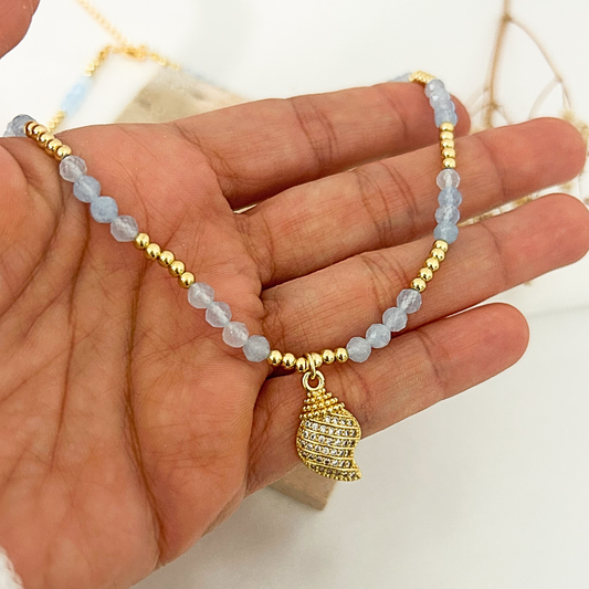 A gold beaded necklace with light blue faceted beads and a gold shell-shaped pendant, shown draped over a hand.