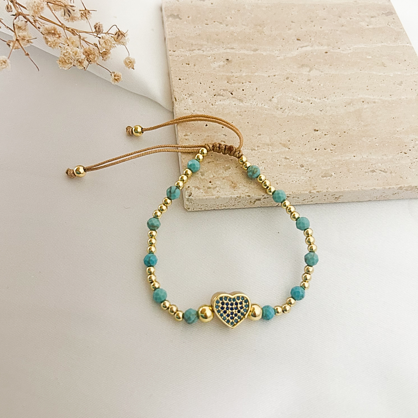 A beaded bracelet with turquoise-blue round beads, small gold spacer beads, and a central gold heart charm set with dark blue stones, displayed on a light surface next to a beige stone tile.