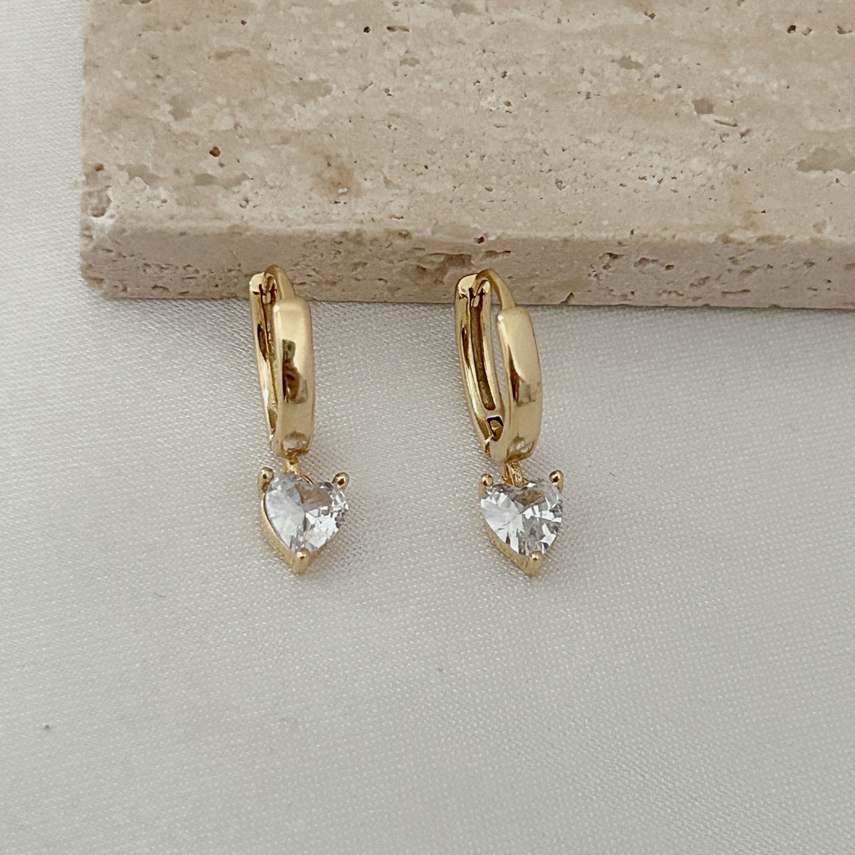 A pair of gold huggie hoop earrings with dangling clear heart-shaped gemstones, displayed on a light beige textured surface with a stone slab in the background.