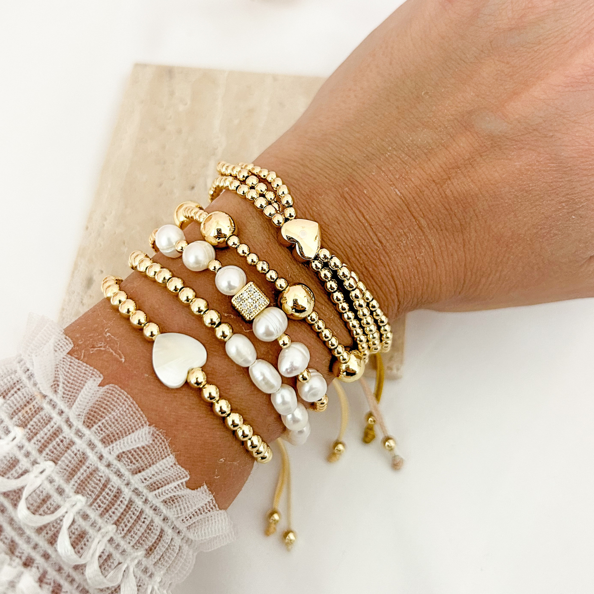 Gold and pearl bracelet stack featuring heart charms, polished beads, and a sparkling square accent.
