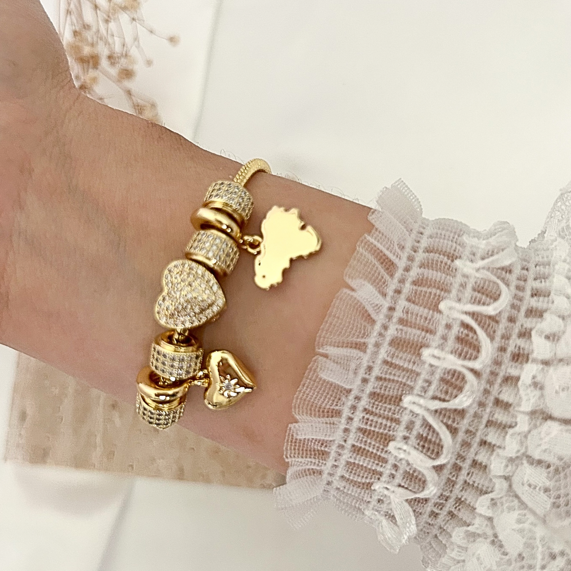 Elegant gold bracelet with crystal-encrusted beads, heart charms, and a polished Venezuela silhouette charm, worn on the wrist with a delicate lace sleeve.