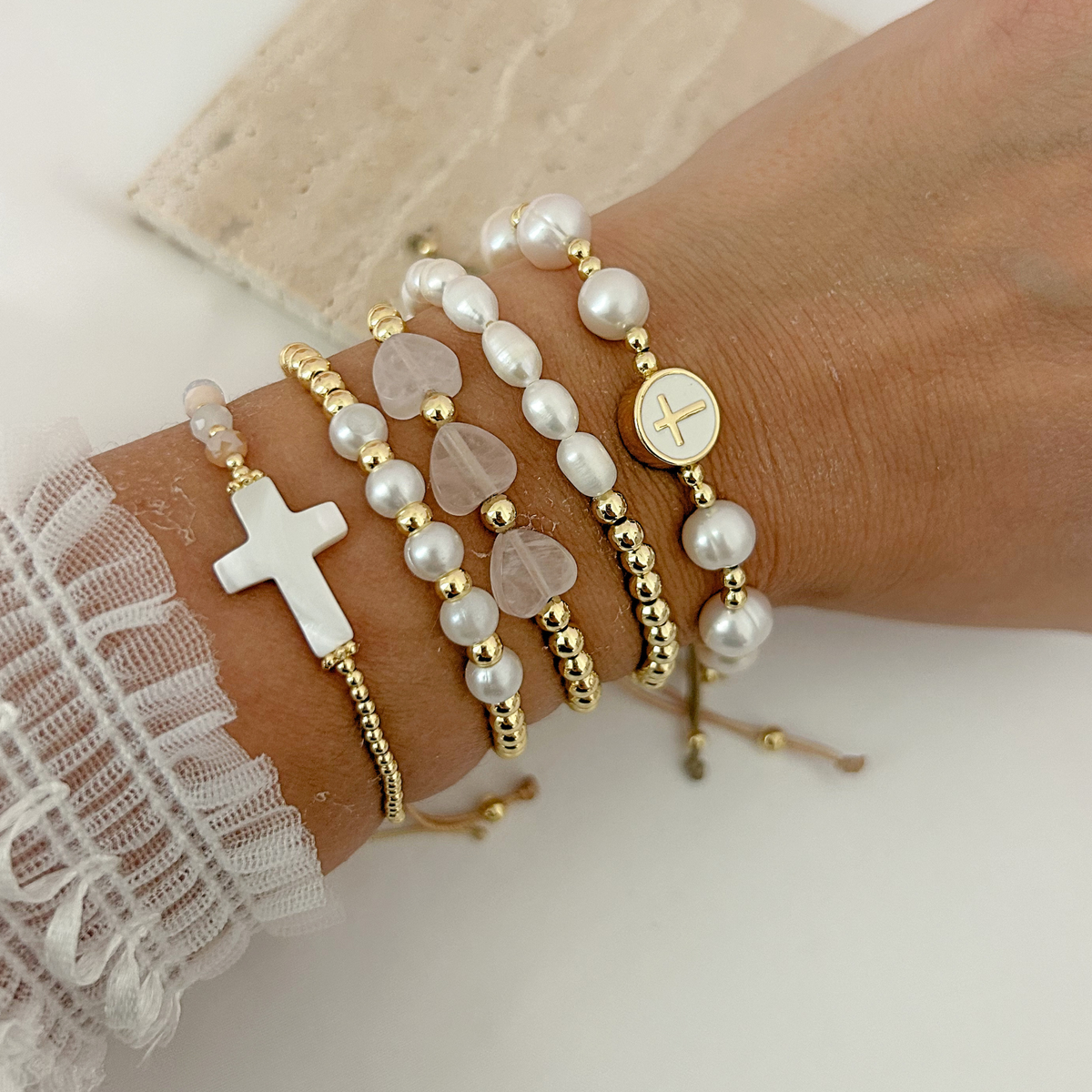 Gold and pearl bracelet stack featuring mother-of-pearl crosses and soft pink heart-shaped beads.